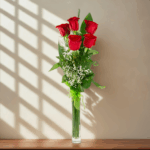 Seis rosas rojas con follaje verde y flores blancas pequeñas en un jarrón de vidrio alto y estrecho sobre una repisa de madera