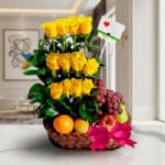 Arreglo floral con rosas amarillas, uvas rojas, manzana, pera y naranja en una cesta de mimbre con lazo rosa, sobre mármol.