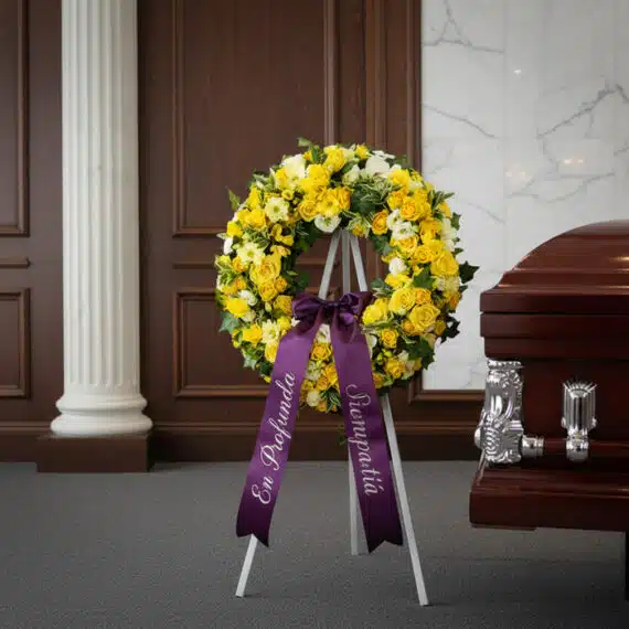 Corona fúnebre de flores amarillas y blancas con cinta morada "En Profunda Simpatía" junto a un ataúd en sala de velación ele