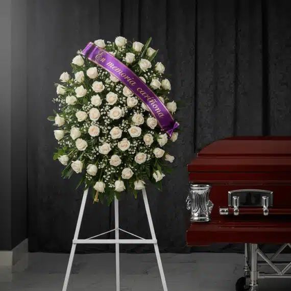 Corona fúnebre de rosas blancas y follaje verde, con una cinta morada que dice 'En memoria cariñosa', junto a un ataúd de mad