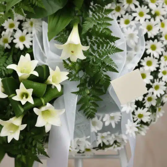 Detalle macro de corona funeraria con lirios blancos, margaritas frescas y follaje verde, con cinta satinada y rocío sutil, p