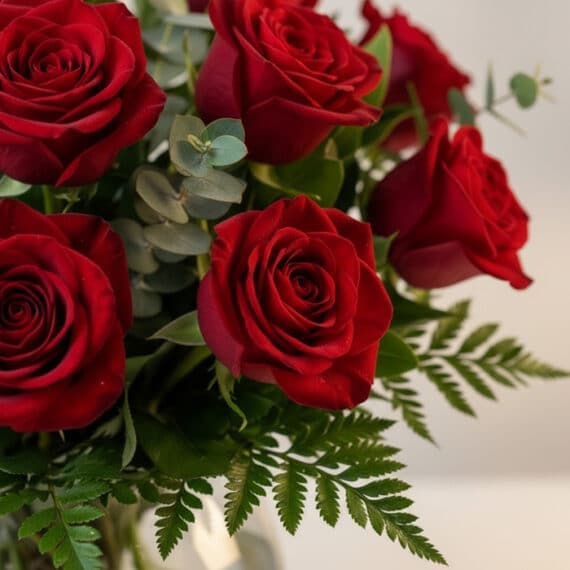 Primer plano extremo de un arreglo floral Jarron Floral Graziella con 12 rosas rojas frescas, gotas de rocío y follaje verde.