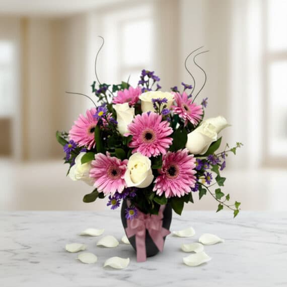 Jarrón Primavera con un elegante arreglo floral de rosas blancas, gerberas rosas y pequeñas flores moradas, sobre una mesa de