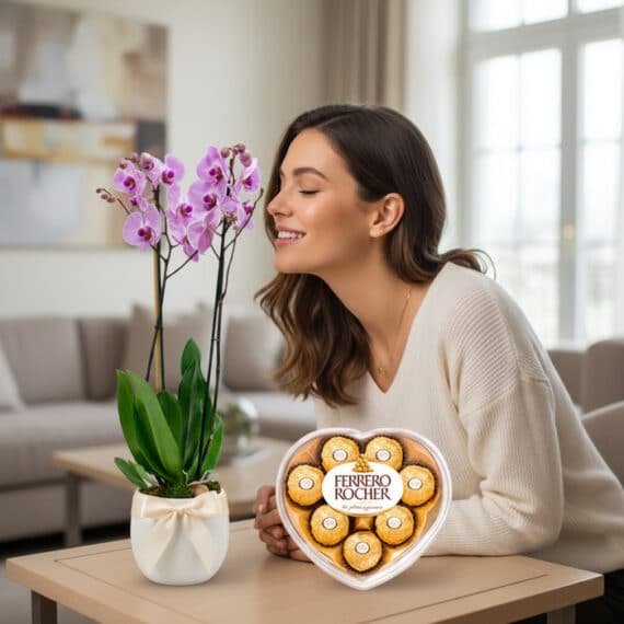 Mujer sonriendo y oliendo una orquídea GALA morada en maceta blanca con lazo, junto a una caja de chocolates Ferrero Rocher e